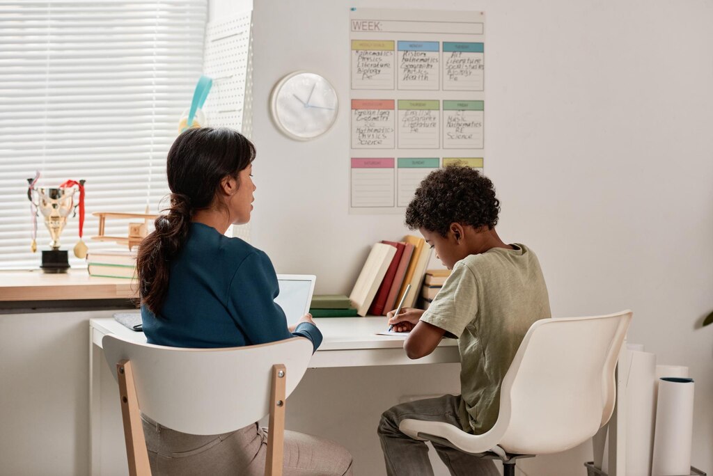 Curlyhaired black boy sitting at table in his room with timetable on wall and doing school task unde